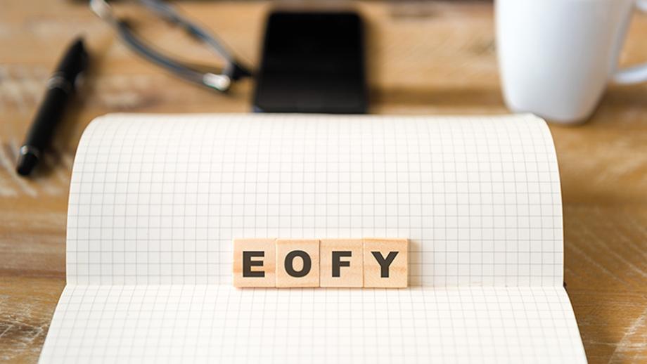a wooden table with a grid-lined notebook open to a blank page. On the notebook are four wooden letter tiles spelling out “EOFY,”